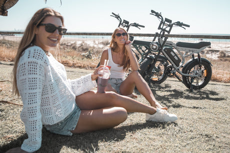 Two women sitting on the ground with electric motorcycles in the background.