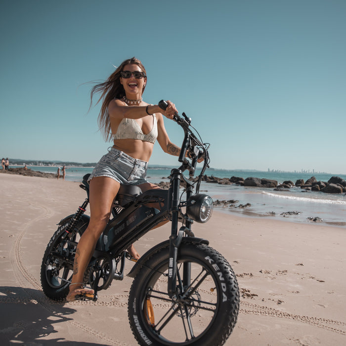 Woman on a beach with an electric bike, wearing sunglasses and a white top.