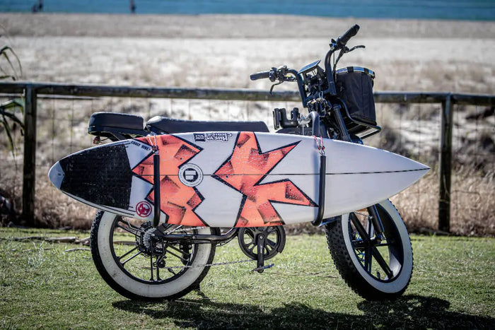Electric Bike with a surfboard rack attached on a grassy area near a beach.
